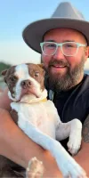 Tim holding his dog while on a beach wearing a hat and smiling